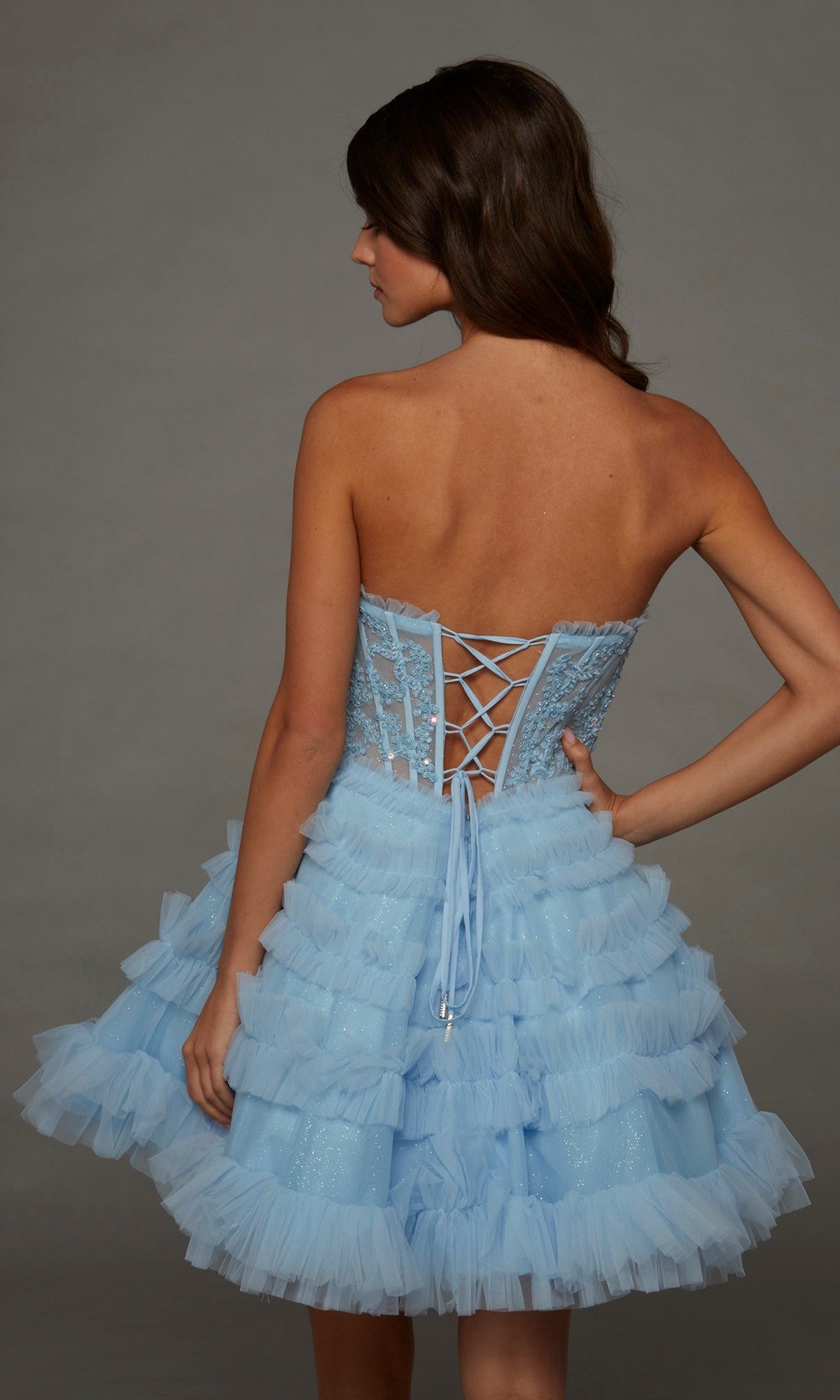 A woman in a strapless, light blue Alyce 30094 short homecoming dress with a lace corset bodice and ruffled skirt stands facing away against a plain gray background.
